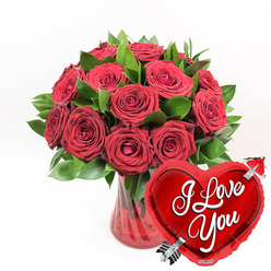 This image captures the essence of a romantic gift you might send to someone special in Grove Park, with every detail crafted to speak of love and thoughtfulness. At its core is a lush bouquet of deep red roses, gathered in a rounded, generous cluster. Around a dozen or more blooms are visible, their velvety petals unfurling in rich layers of crimson. Some roses are fully open, revealing their intricate centres, while others remain slightly cupped, creating a dynamic mix of shapes and textures that feels both natural and expertly arranged. Nestled among the blooms is a collar of fresh, vibrant green foliage, the leaves smooth and gently arching. This greenery frames the roses and adds a crisp contrast that makes the red tones glow more vividly, reminiscent of the vibrant colours seen in local gardens near Grove Park and Gunnersbury. The stems of the bouquet are gathered into a clear, ruby-red glass vase with subtle fluted lines running vertically along its sides. These delicate ridges catch the light, creating soft reflections and giving the vase a sense of movement and refinement. The red tint of the glass echoes the colour of the petals, unifying the overall palette into a warm, romantic harmony. Resting in the lower right foreground, and slightly overlapping the vase, is a gleaming heart-shaped foil balloon in bright, metallic red. Its surface is smooth and glossy, with highlights and reflections that give it a playful, festive feel. Across the centre of the heart, in flowing white script, the words "I Love You" are clearly written. A silver arrow, evoking Cupid's arrow of legend, appears to pierce the heart diagonally from bottom left to top right. Against a pristine white background, these elements combine to form a striking image of devotion, perfect for anniversaries, Valentine's surprises, or any occasion when you want a floral gift to speak clearly of your feelings in Grove Park.