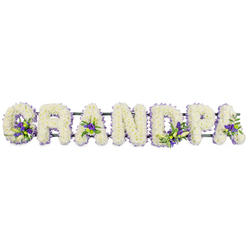 This image captures a refined floral lettering tribute spelling out GRANDPA in substantial block capitals, arranged in a steady horizontal line against a bright white backdrop, evoking the calm setting of a Grove Park farewell. Each letter is entirely covered with tightly clustered white flowers, likely chrysanthemums or similar small-headed blooms, chosen for their durability and their timeless association with remembrance. The mass of petals creates a thick, velvety texture, with hints of shadow between blooms that lend depth and a soft, cloud-like effect. Threaded through this serene white base are clusters of purple and lavender blossoms, interspersed with fresh green foliage that adds a natural, garden-like feel. These accents are strategically placed: in the curve of the G, at the heart of the first A, along the line of the D, around the P, and subtly enhancing the final A. Each cluster is small yet expressive, giving the tribute delicate points of interest while keeping the overall mood gentle and respectful. A slender, dark green framework supports the letters from beneath, nearly invisible but essential in holding the composition together as one continuous tribute. The arrangement feels deeply personal, ideal for placing at the front of a chapel near Grove Park station, by the altar of a local church, or beside a memorial table at a wake along the river. It speaks of shared family stories, Sunday walks through nearby parks, and the steady presence of a grandfather whose life is being honoured with care and artistry.