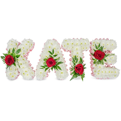 Set against a pure white background, this handcrafted memorial spells KATE in solid, sculpted letters made from countless small white chrysanthemums, their tight heads forming a voluminous, cloud-like surface that reads both tender and formal. Each letter's contour is trimmed with a soft, pale pink ribbon that has been pleated and pinned to follow the edges, adding a subtle warmth and a tailored finish that catches morning light without overwhelming the floral tone.

A single vivid red rose adorns the face of each letter, framed by a generous spray of deep green foliage - a thoughtful mix of broad leaves and delicate fern fronds that create depth and a gentle, natural rhythm. The roses are deliberately positioned: the K's rose sits on the lower section of its left vertical stroke; the A's sits at the apex of its triangular peak; the T's rose is placed at the base of the left side of its crossbar; and the E's rose rests on the central horizontal bar. The combination of pure white blooms, crimson focal roses, and lush greenery conveys a message of love, purity, and remembrance. As made by Grove Park Florist, this personalised tribute is the kind we deliver across Grove Park and neighbouring areas for funerals, wakes, and celebration-of-life ceremonies, created to give families a clear, elegant name tribute that feels both personal and dignified.
