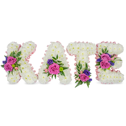 Here the name KATE is shaped into four freestanding letters, each lovingly formed from a thick, plush carpet of pure white chrysanthemums that create a soft, cloud-like surface. The letters are edged with a delicate light-pink satin ribbon that outlines both outer and inner shapes, adding a whisper of warmth against the snowy blooms. Clusters of accent flowers break the white expanse with lively colour: velvety deep-fuchsia roses and smaller pastel-pink spray roses provide the feeling of heartfelt presence, while touches of dark purple or blue statice or lisianthus introduce a quiet, reflective tone. On the K a decorative grouping sits along the diagonal, the A carries a spray near its lower right curve, the T is grounded with a cluster at its base, and the E shows two distinct groupings on the middle and lower bars-each composition carefully balanced to read clearly from a distance. The clusters are cushioned by mixed foliage-delicate fern fronds, soft eucalyptus leaves and glossy greenery-that add depth, scent and a gentle rustle. Bathed in soft, diffused light, the interplay of crisp white, blushing pink, vivid fuchsia and rich purple becomes both visually arresting and soothing. The aroma is quietly floral, driven by fresh roses and green foliage, appropriate for a funeral service, graveside tribute or celebration of life. As a local tribute crafted with neighbourhood care, Grove Park Florist often prepares this personalised style for families attending services near Grove Park station, Chinbrook Meadows or the quiet lanes around Baring Road, with delivery across Grove Park and neighbouring streets when needed.