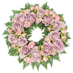 A softly radiant memorial wreath is displayed flat against a clean white backdrop, its perfect circular form filled so completely with flowers and foliage that no gaps interrupt the flow. At its heart, large roses in unique tones of dusty lavender and antique mauve create a sophisticated foundation. Their petals are fully open and plentiful, folding over each other like layers of silk, forming gentle swirls that lead the eye around the ring. Between them, dainty blush pink spray roses and small rosebuds are tucked into every available space, some tightly furled, others just beginning to unfold. This mix of bloom stages adds a sense of movement and time, echoing the many seasons of a life remembered by family and friends in Grove Park. The colour palette is soft and restrained, with the cooler lavender shades balanced by the warmth of the pinks, creating an atmosphere of tenderness rather than stark sorrow. Surrounding the flowers, a thick border of deep green fern fronds fans outward, their feathered, serrated edges creating a lively contrast to the smooth rose petals. The ferns provide structure and a natural frame, their rich green hue enhancing the pastel tones at the wreath's centre. Even, diffused lighting ensures that every detail-from the satin-like roses to the fine texture of the greenery-is clearly defined without harsh shadows. The overall impression is one of tranquil dignity, the kind of wreath that might be chosen for a funeral or memorial near Grove Park station or Chiswick, where families gather to honour a loved one with something beautiful, calm, and deeply respectful.