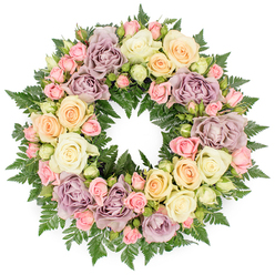 A lush, full funeral wreath is displayed against a clean white backdrop, its perfectly round form composed of closely woven roses and greenery that create an impression of quiet, steadfast love. The arrangement has the slightly formal grace you might expect from a carefully chosen tribute for a service in Grove Park, where families gather in familiar churches and chapels. Dominating the design are generous roses in soft ivory and creamy white, fully opened so that their velvety petals form wide, layered rosettes. Between these larger blooms nestle clusters of blush pink spray roses, some still enclosed as tight buds, others beginning to open, giving the wreath fine detail and a sense of life still unfolding. Gentle touches of pale peach and buttery yellow roses add an inviting warmth, while dusty lavender and muted mauve roses introduce cooler, more reflective tones, lending an air of quiet sophistication. Beneath and around the flowers, dark green fern fronds create a dense, feathery base that both supports and frames the pastel tones, their rich colour bringing the lighter hues into sharper focus. The circular composition symbolises continuity and eternity, appropriate for commemorating a life well lived. Each stem appears thoughtfully placed, as though a florist from Grove Park Florist had taken time to balance colour, texture, and form for a family saying goodbye. The result is a wreath that feels calm, respectful, and deeply compassionate, a floral expression of sympathy that would sit with gentle dignity beside a coffin or memorial photograph.
