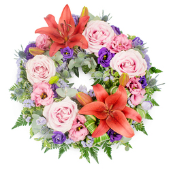 Photographed from above against a bright white background, this circular sympathy wreath appears calm and composed, a ring of colour and texture designed to bring quiet comfort. The wreath's structure is created from a dense layer of greenery, with soft, silvery eucalyptus, feathery ferns, and deep green leaves woven together to form a sturdy, natural-looking base. On top of this foliage, numerous blush pink roses open in gentle spirals, their velvety petals suggesting softness and care, much like a thoughtful gesture offered to a neighbour in Grove Park during difficult times. Threaded between the roses, lisianthus blooms in pale pink and deep, velvety purple add graceful ruffles and a touch of drama, their gently cupped shapes giving a sense of movement as they turn in different directions. Two bold Asiatic lilies in a glowing coral-orange shade are positioned at opposite points on the wreath, their star-like faces and pronounced stamens drawing the eye and symbolising warmth and remembrance. Small clusters of blue-grey eryngium thistles punctuate the design with a spiky, architectural feel, while clouds of tiny lavender-blue filler flowers soften the overall effect, like a delicate haze. Several closed lily buds are nestled among the foliage, hinting at blossoms yet to unfold. Carefully folded and looped green leaves form subtle accents at the bases of the lilies, showcasing skilled florist techniques. The arrangement feels balanced and reflective, ideal as a funeral or memorial tribute for services in and around Grove Park, where its gentle colours and textures can offer solace to family and friends gathered to say farewell.