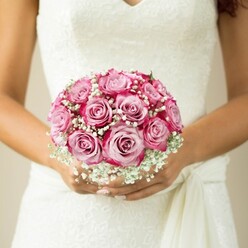 There's an air of calm celebration as a bride, her hands glowing with a natural tan, draws focus to a bouquet that seems almost to float before her lace-adorned dress. Centred on about a dozen vibrant two-tone pink roses, the bouquet blooms with colour-each petal shifting from creamy blush at its heart to vivid magenta at its rim, reminiscent of rose gardens thriving near Chiswick rail embankments or the heart of Grove Park itself. Wispy baby's breath, like puffs of soft cloud, nestles among the blooms, softening the arrangement and bringing an ethereal charm that complements the classical wedding setting. The lace V-neck dress evokes timeless romance, its elegant details mirrored in the playful fullness of the bouquet. With a flowing tie just above her hands, the bride's outfit adds another gentle texture, highlighting her anticipation and delight. The serene, pale background keeps the moment intimate, drawing the observer into a world where expert local florists-like those at Grove Park Florist-make dreams tangible for each unique wedding day in Grove Park.