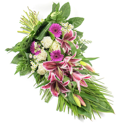 A thoughtfully crafted sympathy spray lies visually against a pure white background, its elongated, diagonal line drawing the eye from the bound stems at the upper left down to a gentle point at the lower right. In the centre of this flowing design, show-stopping Stargazer lilies bloom in deep fuchsia-pink, each star-shaped flower edged in clean white and sprinkled with burgundy speckles, creating a sense of depth and emotion. Some lilies are fully open, while others are still in bud, the closed petals tinged with green and rose, suggesting memories that continue to unfold. Around them, creamy white roses nestle closely, mostly in bud and half-open stages, their smooth, spiral petals bringing a calm, dignified presence. White carnations, with their fine, ruffled edges, fill the spaces between roses and lilies, creating a soft foundation that feels almost cloud-like. Bright magenta gerbera daisies add splashes of vivid colour, symbolising affection and cherished times shared. Tiny white daisy-like filler flowers thread delicately throughout the composition, adding texture and a sense of lightness. All of this is framed by a generous sweep of greenery: broad, glossy dark green leaves lend structure, while airy fern fronds and long, narrow blades of foliage extend outwards and downwards, giving the arrangement its elegant, cascading form. The overall impression is one of solemn beauty and quiet strength, as if prepared in a Grove Park Florist studio just off the main road, ready to be carried to a local church or crematorium. It feels perfectly suited to those moments when words fall short, but flowers can still convey deep sympathy and love.