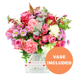 The photograph showcases a romantic mixed bouquet glowing against a pristine white backdrop, every detail sharply defined. Standing in a clean, square glass vase, the arrangement feels both modern and abundantly soft, built around an array of pink roses and carnations. Deep fuchsia and magenta roses nestle alongside gentler blush and pastel pink varieties, their layered petals forming plush rosettes that seem almost velvety to the touch. Between them, frilled carnations in coral, salmon, and peach-pink tones expand the colour palette, their ruffles lending the bouquet a cloud-like, sumptuous texture. Peppered throughout the design are smaller daisy-style chrysanthemums, their petals tinted in lavender and light purple with striking lime-green centres, which brighten the interior of the arrangement and offer a playful contrast. Creamy white blooms are dotted here and there, functioning almost like highlights in a painting, preventing the composition from becoming too saturated with pink. The greenery encasing the flowers is thoughtfully chosen: soft, rounded eucalyptus leaves with a silvery sheen combine with deeper green foliage and a scattering of reddish-brown leaves, bringing depth, structure, and a hint of seasonal warmth. The stems, visible through the crystal-clear glass, are gathered firmly yet gracefully. Around the base of the vase, a neat white band features a repeating motif of tiny red hearts and the phrase all my love written in cursive red lettering, underscoring the bouquet's affectionate message. In the bottom right of the image, an orange circular overlay with strong white capitals announces VASE INCLUDED, reassuring anyone imagining this display in a Grove Park flat or riverside home that it arrives complete and ready to enjoy, perfect for surprise deliveries, birthdays, or simply sending love across SE12.