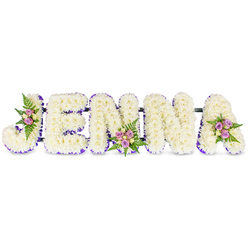 There is a quiet artistry to this personalized floral name tribute: JENNA spelled in five three-dimensional block letters entirely formed from small, creamy white chrysanthemums that give each letter a soft, pillowy texture. The flowers are arranged with exacting care so the letterforms read clearly from across a room, the plush surfaces glowing in subdued, chapel-like light. A slim border of purple - a ribbon or tiny lilac florets - traces the inner edges of every character, offering a gentle line of colour that frames and defines the pristine white. Thoughtful accent clusters appear at three points of the piece: tucked into the J's sweep, settling into the base of the first N, and crowning the upper arm of the A. These mini bouquets combine blush-pink and lavender spray roses, a few pale cream buds, and soft, dark-green fern fronds, bringing a subtle warmth, a faint rose perfume, and natural depth to the overall composition. Behind the letters, slender dark connecting bars hold the arrangement together, a discreet engineering touch that keeps the design steady when placed on a chapel dais or grave side. Perfect for a funeral or memorial service, this tribute offers a personalised, compassionate way to honour a life. Locally made for families in Grove Park and nearby Lewisham neighbourhoods, the piece reflects traditional sympathy floristry - calm, respectful, and crafted for ease of placement in service settings.