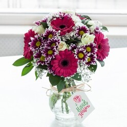 In this image, a professionally arranged bouquet glows against a softly diffused white backdrop, like it has been placed on a bright windowsill in a Grove Park townhouse on a clear morning. The flowers gather in a compact, rounded dome above a clear, subtly textured glass vase, where neatly cut stems can be seen criss-crossing through the water. Dominant among the blooms are generous gerbera daisies in intense shades of magenta and deep fuchsia, each flower defined by its large, flat petals and dark central eye, giving the design an upbeat, modern energy. Interspersed between them are bi-colour chrysanthemums, their many slender petals brushed with rich purple at the tips and fading to clean white toward the centres, which hold small, bright yellow discs. These create a playful contrast of colour and texture. Creamy white roses, both newly opening and still held in bud, introduce softness and a more traditional romantic charm, while airy sprays of white baby's breath weave through the spaces, offering a misty, almost sparkling lightness. Deep green foliage, carefully trimmed and placed, anchors the display and frames the brighter flowers without ever overwhelming them. The vase itself is tied around the neck with a loop of rustic twine, finished in a simple bow, from which a neat rectangular tag with a minimal floral emblem hangs, suggesting a handwritten message tucked in for the recipient. The entire arrangement feels joyful and polished, perfectly suited to local delivery for birthdays, congratulations, or just-because surprises across Grove Park and the nearby streets leading towards Chiswick and Lewisham.