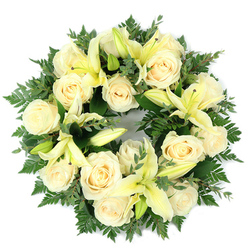 Arranged flat and photographed from overhead, this funeral wreath combines creamy white roses and pale yellow lilies with a lush backdrop of deep green foliage to create a serene, balanced circle of remembrance. Approximately a dozen large roses, their petals tightly swirled and slightly luminous, form the principal band of soft ivory, while lemon-hued trumpet lilies-some fully open, others in bud-offer graceful arcs and a varied vertical rhythm. The greenery beneath includes fine, feathery fern fronds that feather the wreath's edge and broader, rounded eucalyptus-like leaves that introduce a muted, silvery-green tone and visual depth. The contrasts of texture-velvet rose petals, smooth lily petals, and airy fern-are arranged with dense, careful placement so the wreath reads as a single, composed whole. In cool morning or chapel light the blooms take on a tender clarity, their colours suggesting solace and purity rather than spectacle. The scent is imagined as a subtle blend: the soft sweetness of roses balanced by a fresh, almost citrusy green note from the lilies and foliage. As a local tribute, this wreath is the kind of piece we prepare for services across Grove Park and nearby chapels and cemeteries, and it can be arranged for thoughtful delivery by Grove Park Florist when relatives prefer a dignified, comforting gesture sent with care.