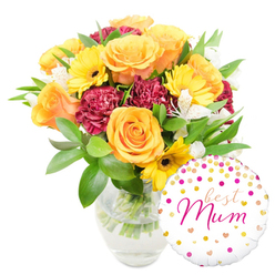 This image presents a thoughtfully composed Mother's Day gift set that reads as both cheerful and expertly arranged. Dominating the left of the frame are sumptuous orange and yellow roses, their petals unfolding in layered spirals that catch soft studio light and suggest a warm, citrus-tinged scent. Bright yellow gerbera daisies intersperse among the roses like small suns, their dark brown centres giving the arrangement visual rhythm, while rich maroon carnations contribute dense, velvety texture and romantic weight. Slender white alstroemeria threads delicately between the bolder blooms, its fine speckling and narrow petals introducing an elegant counterpoint. A lively assortment of deep green foliage-glossy leaves and feathery sprigs-wraps the composition, offering both structure and a fresh, earthy contrast. The stems rest in a clear glass vase filled with water, the transparency underscoring the bouquet's newly-arranged quality and careful preparation. Nestled to the lower right, partly overlapping the vase, a round white foil balloon decorated with gold and hot-pink polka dots and tiny hearts proclaims "best Mum" in flowing hot-pink script, giving the image an unmistakable celebratory focus. As a local florist serving Grove Park, Lee and Hither Green, Grove Park Florist often pairs blooms and balloons like this for mothers who live near Chinbrook Meadows or need a thoughtful doorstep surprise; the photo conveys the tactile softness and joyful intention behind such a gift without losing the quiet craftsmanship of the arrangement.