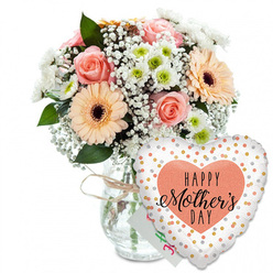 This photograph captures a warm and joyful Mother's Day gift arrangement styled with understated elegance. The scene is set against a spotless white background, which makes every colour appear clear and bright, as if the bouquet were resting on a sunlit table in a Grove Park home. A clear glass vase stands in the centre, its straightforward, cylindrical form giving a clean, modern base for the flowers above. A slim wrapping of rustic twine encircles the neck, forming a small, informal bow that adds a hint of handmade charm. Emerging from the vase, the bouquet forms a soft, rounded dome, filled with pastel tones and crisp whites. Fully opened peach-pink roses reveal tight spirals of velvety petals, and their gentle blush tones are echoed in the peach and light orange gerbera daisies positioned around them. These gerberas have broad, layered petals framing distinctive dark brown centres, providing a focal point within the arrangement. Between the larger blooms, airy clusters of white baby's breath give a subtle mist-like effect, while compact white chrysanthemums with apple-green centres bring structure and a fresh, contemporary note. Deep green foliage is visible throughout, framing the pastel flowers and evoking the leafy character of Grove Park's gardens and riverside paths. Slightly obscured behind the blooms, a small white card peeks through, its partially visible green and pink lettering implying a handwritten Happy Mother's Day message. To the right of the vase, leaning forward, a generous heart-shaped foil balloon glows softly. Its bright white background is scattered with tiny metallic gold and silver dots, and at its heart lies a peach-coloured shape bearing the phrase HAPPY Mother's DAY in bold black text, with an elegant flourish to the word Mother's. The composition radiates affection and appreciation, as if ready to be carried down a Grove Park street to surprise a much-loved mum.