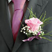 This image shows a sharply dressed guest or groom, captured from the shoulders down, standing ready for a celebration somewhere around Grove Park's quiet residential lanes. He wears a dark charcoal or black suit jacket, the fabric smooth and well-fitted, with a subtle sheen along the lapel where the light falls. At the center of the frame, pinned securely to the left lapel, is a romantic boutonniere built around a single pale pink rose. The rose is at full, flawless bloom, its velvety petals curling outward from a richer, warm pink core to lighter, blush-toned tips, like a soft sunrise against the dark suit. Encircling the rose, clusters of white baby's breath form tiny, airy dots, giving the arrangement a gentle, misty texture, almost like a miniature cloud resting on the jacket. Slender green fronds of foliage stretch up behind the flowers, lending lift and a natural, meadow-fresh feel, as though the rose has just been gathered from a garden. The stems are bound tidily with a deep green wrap, finished with a delicate satin bow in muted mauve or lavender, its sheen catching the light and subtly tying in with the smooth, fuchsia-magenta tie beneath. The tie lies neatly over a light dress shirt with a soft checked pattern, adding quiet structure without overpowering the florals. The composition suggests a calm, anticipatory moment before a wedding or prom, perhaps after a florist from Grove Park has just pinned the piece in place, ensuring every stem sits perfectly. The atmosphere is affectionate and celebratory, capturing new beginnings, heartfelt promises, and the thoughtful grace of a well-chosen floral detail.