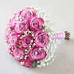 This photograph captures an elegant hand-tied bouquet resting on a soft, linen-like backdrop in a warm, light beige tone, much like the calm interiors of homes around Grove Park. The bouquet is composed of approximately twelve roses, each one a carefully layered gradient of colour: the inner petals hold a muted, dusty mauve or lavender-pink, gradually deepening as they spiral outward into bold fuchsia and deep magenta. The roses are in tight to semi-open bloom, showing off intricate curls and folds that create a sense of richness and depth. Cradling these central blooms is a generous cloud of white baby's breath, whose countless tiny gypsophila flowers form a light, airy frame, softening the dramatic pinks and giving the arrangement an almost ethereal glow, like mist hanging above Duke's Meadows early in the morning. Here and there, small portions of deep green leaves peek from beneath the mass of petals and gypsophila, adding a touch of natural contrast. The stems of the bouquet extend diagonally toward the bottom right and are bound together in a sleek lilac satin ribbon, its smooth surface catching the light with a gentle shine. Encircling this ribbon is a slender string of white pearls, winding in a delicate spiral and adding a sense of classic bridal elegance and formality. The overall mood is romantic and poised, perfect for a wedding at a local Grove Park venue, a bridal shower, or a special anniversary celebration at home. The artistry in the placement of each rose and the careful wrapping of the stems reflects the work of an experienced florist who understands how to blend colour, texture, and sentiment into a single, memorable bouquet.