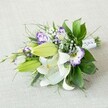 An elegant hand-tied bouquet is laid diagonally across a softly textured, light beige fabric, the neutral background allowing every bloom to stand out with quiet confidence. At the heart of the arrangement, several large white lilies are fully open, their velvety petals curling back to reveal golden-yellow stamens dusted with pollen and a scattering of fine reddish-brown speckles near the throat. Around them, plump chartreuse-green buds stretch along the line of the bouquet, promising more lilies to open in the days ahead, the sense of unfolding beauty echoing the calm, leafy streets of Grove Park. Threaded between the lilies are ruffled Lisianthus flowers, each bloom a little cup of white edged in rich purple, some leaning towards soft lavender, others deep violet, creating a painterly play of colour. Tiny clusters of white baby's breath hover like delicate mist, softening the transitions between the stronger shapes and hues. A generous collar of foliage surrounds and supports the flowers: glossy, dark green leaves provide structure and depth, while finer, more feathery greens extend outwards, adding movement and an almost windswept grace, as if touched by a gentle breeze drifting up from Chiswick or the nearby river. The stems are neatly gathered and wrapped in smooth white satin ribbon, subtly accented with tiny pearl-like beads that catch the light, hinting this could be a bridal bouquet or special celebration piece carried into a Grove Park ceremony. The lighting is gentle and diffused, with no harsh shadows, highlighting the freshness of the petals and the lushness of the greenery. The overall impression is one of purity, romance, and quietly luxurious craftsmanship, the sort of bouquet a local florist might prepare for a wedding morning or an intimate anniversary dinner at home.