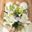 On a joyful wedding day in Grove Park, a bride holds close a bouquet that is as evocative as a gentle breeze along the Thames. At the center, radiant white Oriental lilies reveal their full, fragrant petals, some gracefully speckled and all exuding a quiet purity. Tucked between are clusters of lisianthus, their tender ruffles edged with whimsical lavender, and a few still cocooned, ready to bloom in their own time. Striking purple Veronica, reminiscent of springtime walks through local parks, adds flair and drama in slender peaks. Delicate baby's breath wreaths the arrangement with its cloudlike softness, blending seamlessly with abundant fresh green foliage-unopened lily buds, bold broad leaves, and feathery asparagus fern that spill playfully to the edges. The bride's perfectly manicured hands, topped with subtle polish, cradle the bouquet's stems, the soft, blurred backdrop of a white strapless dress lending dreamy contrast. This bouquet is more than a floral creation; it's a tactile expression of Grove Park's neighbourhood spirit-filled with anticipation, love, and the serene, organic beauty that defines special celebrations near Grove Park's leafy avenues.