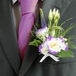 A close-up wedding portrait focuses on the upper torso of a man dressed in refined formalwear, standing as if ready to greet guests arriving through the leafy streets of Grove Park. He wears a dark charcoal grey suit jacket, perfectly tailored so the shoulders sit cleanly and the lapels lie flat and confident. Beneath the jacket, a light grey checkered dress shirt adds a quiet pattern, its crisp cotton catching the soft indoor light. At the centre of the frame, a lustrous magenta silk tie is knotted with care, its smooth sheen glowing warmly against the cooler tones of the suit. Pinned to the left lapel is a meticulously crafted boutonniere: at its heart, two white lisianthus blooms edged in vivid purple, their gently ruffled petals encircling sunny yellow centres that bring a delicate brightness. Around them cluster slender, unopened white buds, suggesting new beginnings and the promise of the day ahead. A single, upright spike of deep indigo-purple veronica gives height and a graceful line, while tiny white filler flowers, much like baby's breath, nestle between tufts of feathery greenery and glossy emerald leaves. Fine, looping strands of decorative green grass add a subtle contemporary twist, curving lightly around the flowers. At the base, a neat white satin ribbon bow gathers the stems, its soft sheen echoing the polish of the tie. The overall impression is one of composed joy and quiet anticipation, the kind of thoughtfully coordinated groom's detail a local Grove Park florist might create for an intimate ceremony in a nearby church or garden venue.