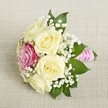 A chic, compact bouquet rests on a creamy, textured linen background, its artistry echoing the refined aesthetics of Grove Park Florist. The arrangement centres around five luminous, cream-coloured roses, each petal unfurling smoothly and fresh-a quiet nod to the riverside elegance of Grove Park. Discreetly nestled off-centre is a single bold magenta rose, injecting a lively splash of colour amid the pale blooms and hinting at joyful celebrations. Sprigs of delicate white baby's breath weave through the bouquet, imparting an airy softness and enhancing its timeless feel. Accents of deep green foliage anchor the design, adding natural vibrancy. The stems are elegantly wrapped in a bright pink satin ribbon, adorned with a spiralled trim of pearl beads, bringing modern sophistication to a classic form. Gentle, diffused lighting highlights the intricate floral textures, evoking the charm of Grove Park's sunlit mornings. This bouquet, ideal for a bridesmaid or a cherished moment at a wedding venue near Chiswick Station, combines romance and expert local floristry for a result that is both memorable and beautifully personal.
