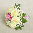 A delicately crafted petite bouquet rests diagonally across a neutral, light beige fabric that looks like finely woven linen, its gentle texture framing the flowers without distraction. At the centre, five creamy white roses, verging on ivory, are arranged in a close cluster, each bloom at that ideal stage between bud and full openness, when the petals are soft, velvety, and slightly cupped. Their subtle, powdery fragrance is almost imaginable from the picture, calm and understated rather than overpowering. Breaking the soft neutrality, a single bright fuchsia-pink rose is set slightly forward on the left-hand side, its richer hue offering a playful, romantic contrast and suggesting individuality-perfect for someone who wants a hint of colour in an otherwise classic design. Wisps of white baby's breath weave throughout the roses, their tiny blooms giving a light, misty effect, like a veil of fine lace around the main flowers. Deep green pointed foliage peeks out from beneath and behind the blossoms, giving structure to the compact arrangement and grounding the softer tones with cool, natural depth. The stems are tightly bound with a smooth, bright pink satin ribbon that catches the light in subtle highlights, forming a polished handle. Over this, narrow rows of small white pearls are arranged with jeweller's precision, adding vintage charm and a sense of occasion. The soft, even lighting and careful composition suggest a bouquet ready for a Grove Park bride, prom guest, or proud parent on a special evening, the kind of thoughtful finishing touch often prepared for same-day delivery by local florists serving the surrounding London streets and parks.