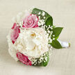 A hand-tied bouquet is laid out horizontally on a light beige, slightly textured fabric, the soft, natural look of the material evoking the relaxed charm of homes and cafes around Grove Park. At its centre, a generous creamy white peony unfurls fully, with layers upon layers of delicate, ruffled petals creating a sumptuous, cloud-like bloom that immediately draws attention. Clustered closely around this focal flower are fuchsia-pink and dusky blush roses, their petals tightly coiled in perfect spirals, giving the arrangement a sense of classic romance and depth. Scattered delicately among the larger blooms are fine sprays of white gypsophila, known as baby's breath, whose tiny, star-shaped flowers create a light, airy veil, softening the transitions and adding a lace-like texture. Encasing the flowers, broad dark green leaves curve gracefully along the base and sides, slightly glossy and sculptural, providing both structure and an appealing contrast against the cream and pink tones. The stems are carefully gathered and wrapped in a smooth white satin ribbon, producing a neat, elongated handle that suggests the bouquet is intended to be carried down the aisle or displayed proudly at a wedding reception near Grove Park or Strand on the Green. Gentle, even lighting washes over the scene, enhancing the subtle shadows in the peony's layers and the soft gradients in the rose petals, while preserving an overall feeling of calm and romance. The bouquet feels fresh, thoughtfully composed, and quietly luxurious-ideal for intimate ceremonies, anniversary surprises, or same-day wedding flower delivery across the Grove Park area.