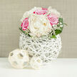 On a clean, bright white tabletop, a lush bouquet of creamy peonies and fuchsia-pink roses becomes the focus, like a carefully placed centerpiece in a Grove Park townhouse near the river. The bouquet is compact and rounded, reminiscent of a bridal posy, with the soft ivory peonies forming the pillowy heart of the design. Each peony is full and open, its layered petals slightly ruffled and luminous in the gentle, even light. Around them, roses in vivid shades of pink add drama and romance, their velvety petals spiralling tightly and deepening in colour at the edges. Delicate sprays of white baby's breath thread through the composition, their tiny blossoms creating a fine, cloud-like texture that bridges the space between the larger blooms. At the base, broad, glossy green leaves curl outward in graceful arcs, framing the flowers and enhancing the domed silhouette. The bouquet is displayed in a distinctive white vase with an open, woven structure, as if slender, painted branches have been intertwined into a decorative sphere. Through the criss-crossing lines, glimpses of stems and shadow add an intriguing sculptural quality. In the foreground sit two pale, rattan-like decorative spheres, one slightly larger than the other, subtly echoing the vase's material and reinforcing the natural, organic theme. The background is a soft, neutral beige, possibly a linen-textured wall or fabric, which allows the colours of the flowers to stand out without distraction. The overall impression is one of gentle sophistication and celebratory calm, perfect for wedding décor or a special floral delivery to mark a milestone in Grove Park.
