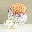 Resting on a sleek white surface that reflects a hint of the arrangement above, this floral centerpiece exudes the calm, polished charm of a wedding table set in a Grove Park townhouse or a nearby riverside venue. The focal point is a flawless dome of roses in peach and apricot tones, their petals soft and velvety, clustered so tightly that they form a perfectly rounded mound of colour. Each rose appears at its peak, open enough to show the delicate spiral of the centre while still holding a gentle, curved form. Around the base of this rose dome, a ring of white lisianthus blooms adds a refined collar. Their petals are ruffled and edged in deep purple and violet, creating an elegant contrast that frames the warm hues above. Between the lisianthus, tiny sprays of white baby's breath are woven like lace, softening the transition from flower to container. The blooms are arranged in a bulbous white woven vase, its shape created by interlacing strands of wicker or rattan that cross over one another in a loose, open lattice. This structure allows shadows and light to move through, adding depth and interest around the base of the flowers. In the foreground, two decorative spheres, also woven from pale, natural-toned material, sit slightly apart on the same surface, one larger and one smaller, echoing the globe-like form of the bouquet. Behind the entire scene, a neutral beige, subtly textured background provides a quiet, warm canvas. The result is a sophisticated, romantic composition, ideal for envisioning wedding flowers or special-occasion décor in Grove Park, crafted with the attentive eye of a local florist who understands the area's relaxed yet elegant style.