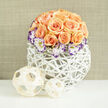 A dense, rounded cluster of roughly two to three dozen apricot and peach roses takes centre stage, their petals layered in concentric rhythm so the arrangement reads as both abundant and carefully restrained. Interwoven at the base are pristine Lisianthus blooms-white with deep indigo or violet edging-that offer a crisp, painterly contrast to the warm rose tones, while hints of Gypsophila pepper the bouquet with delicate, cloud-like accents that soften the overall silhouette. The flowers sit within a modern white orb of woven reeds; the open lattice vase resembles wicker or rattan work and gives the composition an airy, handcrafted feel that plays beautifully against the bouquet's dense texture. Placed on a clean white surface, the display is accompanied to the left by two pale wooden spheres, one smaller than the other, whose delicate weaving mirrors the vessel and adds a casual decorative note. A warm, linen-coloured wall behind is softly blurred, keeping attention on the flowers' colour and form under gentle daylight. As a florist at Grove Park Florist I imagine this piece gracing a local wedding table near Grove Park station or brightening a living room close to Chinbrook Meadows-its scent subtle, its presence refined. The arrangement communicates celebration and tenderness-an elegant gift for anniversaries, a serene centrepiece for intimate events, or a thoughtful gesture to mark new beginnings-all while reflecting the considered craftsmanship and seasonal freshness patrons in the Grove Park area appreciate.
