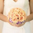 A close-up, chest-level photograph shows a bride standing quietly in her wedding gown, the frame focused on the bouquet resting gently in her hands. She wears a classic white or soft ivory dress with a lace-trimmed bodice and a modest V-neckline, the delicate pattern catching the light in a way that feels both traditional and romantic. The fabric of the skirt falls in soft folds, forming a calm, neutral backdrop that allows the flowers to take centre stage. Nestled between her lightly manicured fingers is a perfectly round bridal bouquet, densely packed with roses in shades of peach, apricot, and blush pink. Each rose is tightly furled, its velvety petals forming spirals that move from pale outer tones to warmer, more saturated hearts, like the glow of a Grove Park sunset over nearby green spaces. Beneath the main rose dome, a striking ring of deep purple blooms edged in creamy white, likely lisianthus, wraps around the base, adding dramatic contrast and a sense of depth. Tiny white filler flowers, similar to baby's breath, peek out in airy clusters, softening the line where the bouquet meets the gown. The bride's nails are painted in a pale, understated shade, with a hint of shimmer on one thumb that mirrors the bouquet's subtle sparkle in the natural daylight. The entire composition suggests calm excitement and tender anticipation, like the moments just before walking into a ceremony at a local Grove Park church or registry office. It feels like a bouquet chosen with care, designed for a bride who wants timeless romance with a modern twist of colour, and captured at the exact instant when love, craft, and quiet joy come together in a single image.