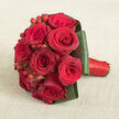 A tightly gathered bouquet of deep red roses lies across a pale beige, fabric-textured surface, like a carefully placed bridal posy resting in a Grove Park home before it is carried down the aisle. The roses are massed into a rounded shape, each bloom brimming with layers of velvety petals that unfold from dark, burgundy shadows into rich crimson highlights. Between these luxurious rose heads, tiny clusters of smooth red hypericum berries are tucked in, their glossy surfaces glinting lightly and bringing a playful sparkle to the otherwise soft, velour-like composition. Encircling the base of the arrangement is a neat, structured collar of dark green leaves, each leaf bent and folded with precision, creating a clean frame that both supports and visually anchors the bouquet. The stems are wrapped firmly in vivid red satin ribbon, its sleek texture catching the light and giving the handle a refined, cohesive finish that feels ideal for a bride stepping out near Grove Park station or a partner planning a surprise anniversary dinner. Captured from a slightly raised viewpoint, the photograph reveals the interplay of textures-the plush petals, polished berries, and smooth foliage-while the neutral beige background withdraws politely, ensuring the romantic reds and greens remain the focal point. The bouquet's mood is intimate and heartfelt, well-suited to wedding celebrations, Valentine's Day gifts, or elegant same-day deliveries arranged by a Grove Park florist for nearby homes along the tree-lined streets by the river.