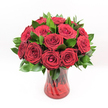 A classic dozen red roses stands proudly against a pure white studio background, every detail of the bouquet quietly magnified by the clean, uncluttered space. Each rose is a deep, velvety crimson, the petals layered like soft silk, unfurling from tight, romantic centres into generous, open blooms. The flowers are arranged in graceful symmetry, facing outward as though ready to meet the gaze of the person they're meant for. Around them, glossy dark green foliage, likely Ruscus, peeks between the roses and fans gently at the base, adding depth, structure, and a natural contrast that makes the reds glow even richer. The bouquet is gathered in a fluted ruby-red glass vase, its translucent sides rippled with a subtle swirling texture that catches the light like reflections on the River Thames on a bright afternoon near Grove Park. Through the tinted glass, the rose stems are visible, crossing and intertwining in darker vertical lines that anchor the arrangement and lend a sense of grounded elegance. The lighting is bright, even, and honest, illuminating every fold of petal and the faint sheen on each leaf, so the flowers look freshly placed-just as they might appear when delivered to a townhouse off Grove Park Road or a cosy flat near Chiswick. There's an implied fragrance in the image: the soft, familiar scent of classic red roses, suggesting love, devotion, and heartfelt emotion. This is an arrangement suited to grand romantic gestures-Valentine's Day surprises, milestone anniversaries, or any moment when words feel too small and only the timeless beauty of red roses will do.