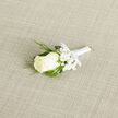 Nestled on a soft, pale beige woven cloth, this boutonniere centres on an immaculate creamy white rosebud whose petals fold inward in delicate symmetry. Tiny sprays of baby's breath form a misty frame, their snow-like points softening the silhouette, while slender, feathery sprigs of dark green-similar to cedar or evergreen-and broader glossy leaves create a layered, verdant backdrop. The stems are bound with a satin-white ribbon, its sheen catching the light and tied into a petite, disciplined bow that speaks of quiet craftsmanship. Laid diagonally, the piece catches a gentle side light that reveals the subtle gradients of ivory and the textural difference between velvety petals and crisp foliage. The fragrance is imagined as a clean whisper of floral sweetness, the kind you notice when stepping into a church or garden on a cool morning. As a local florist from Grove Park, with many bouquets prepared for couples marrying near Grove Park Nature Reserve and guests travelling from Hither Green and Lee, I recognise this boutonniere as a classic choice: simple, symbolic and long-lasting on the lapel. It suggests new beginnings, sincerity, and a respectful nod to tradition, suitable for grooms, ushers, or an honoured guest. The neutral, natural-fibre backdrop enhances the boutonniere's fresh colours and fine details, allowing the viewer to appreciate the florist's attentive hand without distraction. This is an understated, elegant accent that complements formal wear while preserving a soft, intimate mood.