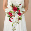 In this intimate wedding portrait, the camera lingers on a breathtaking cascading bouquet, cradled at the bride's waist so that only her arms and the sculpted bodice of her white gown frame the composition. The dress shows subtle lace and embroidery, but it is the flowers that capture the eye: lush crimson roses sit at the centre, each bloom densely petalled and velvety, their deep red tone reminiscent of classic romance on a quiet evening in Grove Park. Nestled among them, pure white lilies unfurl with stately poise, their wide petals forming star-like shapes that reveal bright green stamens at their core, suggesting a gentle, fresh fragrance. Flowing from this focal cluster, elegant white Phalaenopsis orchids cascade in graceful arcs, their smooth, waxy petals and tiny buds trailing downward like a floral waterfall, echoing the gentle curves of the nearby riverbank paths. Wisps of baby's breath meander through the design, creating a light, cloud-like veil that softens the striking red-and-white contrast. Glossy red hypericum-style berries stud the bouquet, catching the light and adding cheerful punctuation marks of colour and texture among the petals. Embracing the whole arrangement is a rich collar of greenery, from fine, ferny fronds to wider, deeper green leaves that lend structure and a sense of movement, like the foliage that lines Grove Park's tree-lined streets. The bouquet forms a classic teardrop silhouette, with each bloom carefully placed to ensure a natural yet deliberate flow. Behind the bride, the background fades into a warm, neutral blur, keeping all attention on this romantically styled red-and-white bridal bouquet, perfect for a heartfelt Grove Park wedding celebration.