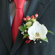 Framed tightly against the left lapel of a well-cut charcoal suit, this boutonnière presents a single luminous Phalaenopsis orchid as its focal point, the white petals smooth and cool, their edges softened by gentle ambient light. At the flower's centre the yellow-gold patterned labellum reads like a delicate, sunlit brushstroke, giving the bloom a quietly radiant core. Complementing this are deep, glossy green leaves that anchor the composition and feathery sprays of gypsophila whose tiny white blooms lend a cloud-like lightness. A neat cluster of pink-to-red hypericum berries nods toward the groom's solid red satin tie, the berries' plump, waxy texture creating an inviting contrast to the orchid's silk. A slender red ribbon encircles the base, its sheen echoing the tie and adding a considered finish. The suit's lapel and a subtle light-blue or grey patterned shirt visible beneath add a sartorial frame that emphasizes the boutonnière's formality and tenderness. As a local florist in Grove Park, I imagine delivering this piece across Grove Park station and the surrounding streets for a wedding, anniversary, or formal evening: its restrained fragrance, textural contrasts and colour harmony make it equally suited for church vows, a civic gala, or a prom photograph taken on Elmstead Lane. The overall impression is one of quiet confidence and careful craftsmanship - a tiny, wearable bouquet composed with the same attention to balance and sentiment we bring to every order from Grove Park Florist.