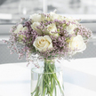 A softly lit sympathy bouquet of white roses and delicate lavender filler flowers stands in a tall glass vase, its calm presence captured in a close, intimate view. The roses are the first thing your eye would find: fully opened blooms with creamy white petals layering one over another in intricate spirals, giving each flower a sense of depth and softness, like folded satin. Threaded between these larger blossoms are fine sprays of tiny lavender and lilac-toned flowers, probably statice, forming a light, cloud-like frame that softens the outline of the bouquet and adds a gentle touch of colour without disturbing its quiet mood. The arrangement is held in a simple, cylindrical vase made of clear glass, allowing the straight green stems to be seen clearly as they reach down into cool, clear water, suggesting freshness and recent, careful arranging. Resting on a smooth, bright white surface, the vase casts subtle reflections, while the surrounding light creates soft highlights and mild shadows across the petals. The background blurs into pale greys and whites, evoking a peaceful, uncluttered room in Grove Park-perhaps a sideboard near a window overlooking one of the area's residential streets or green spaces. The overall feeling is one of gentle support and heartfelt respect, making this bouquet an apt choice for conveying sympathy, whether delivered to a family home or a quiet service nearby. The image holds a sense of stillness and contemplation, like a floral whisper of comfort during a tender time.