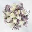 In this overhead view, a round sympathy bouquet sits like a calm, floral circle on a bright white backdrop, its muted tones and soft textures creating a feeling of quiet solace. The central part of the design is formed by approximately a dozen white roses, placed closely together so that their petals intersect and form a gentle, undulating surface. Some roses are still partly closed, with smooth outer petals hugging their centres, while others are more open, revealing intricate spirals of creamy white with a hint of pale ivory hidden at the heart. The petals appear thick, velvety, and cool, suggesting the freshness you'd expect from flowers prepared that morning in Grove Park, ready for delivery to a family gathering or remembrance service. Surrounding and threading through the roses are sprays of lavender-hued Limonium, also known as Statice, their tiny blossoms creating a fine, misty halo of soft purple. This filler adds a delicate, papery texture that contrasts with the plush roses, giving the bouquet depth and a sense of lightness, as though it might float. Subtle touches of green from slender stems and minimal foliage peek out between the blooms but remain secondary to the white and lavender palette. The light is clean and diffused, highlighting every curve and edge without sharp shadows, which enhances the arrangement's serene and contemplative tone. It feels like the kind of design a caring Grove Park florist would assemble for condolence deliveries around the area, from homes near Grove Park station to intimate services along the river, offering a gentle message of sympathy, remembrance, and peace.