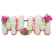 A tender, standing floral tribute spells out the word MUM in large connected block letters, photographed against a clean white backdrop so every detail reads clearly. Each letter is densely built from small, pristine white chrysanthemums whose feathery petals create a soft, cushiony surface and a serene, even texture across the whole message. The outer edges of the letters are finished with a delicate ruffled trim of pale pink ribbon that adds a whisper of warmth and a subtle, motherly softness to the composition. Between the letters a dark green support frame is faintly visible, giving structural stability while remaining unobtrusive. Three small accent posies punctuate the design: a cluster of deep fuchsia-pink roses with glossy ferny foliage and tiny coral-red hypericum berries nestles at the upper left of the first M; a matching spray sits in the base curve of the U, drawing the eye to the centre; and a third cluster decorates the upper right of the final M. The contrasts-crisp white mums against velvety pink roses, soft ribbon against the cool green stems, and the small bursts of berry-red-create a tactile, comforting palette. There is almost a faint, clean floral scent suggested by the mums, tempered by the heady rose perfume if one leans in. This arrangement reads as a calm, respectful expression of love and remembrance for a mother-ideal for a funeral service, memorial or graveside dedication-designed with the quiet dignity a family seeks when saying goodbye.