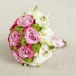 A romantic hand-tied bridal bouquet is photographed from a gently elevated angle, resting on a softly textured light beige fabric that feels like a calm, neutral backdrop to its colour and detail. At the centre, several fully opened roses in soft lavender-pink bloom with satin-like petals that deepen to a richer, almost fuchsia edge, giving each flower a graceful two-tone swirl. Nestled between these roses are compact clusters of bright chartreuse green button chrysanthemums, their tiny, domed heads adding playful texture and a lively freshness that lifts the whole arrangement. Delicate white blooms, likely lisianthus or a similar flower, weave through the design, their layered, ruffled petals and pale green-yellow centres lending a gentle, cloud-like softness. A few tight green buds peek out here and there, suggesting that the bouquet has been freshly arranged, perhaps just prepared for a ceremony in one of Grove Park's local churches or a celebration at home near the station. The stems are neatly gathered and wrapped in smooth pink satin ribbon, forming a comfortable handle that feels luxurious in the hand. Over the ribbon, a slender strand of tiny white pearls spirals down the length of the grip, echoing vintage jewellery and bridal accessories. The soft, even lighting highlights the contrast between the pink, white, and vivid green blooms without casting harsh shadows, giving the bouquet an elegant, timeless presence. It's an arrangement that would suit a Grove Park wedding, anniversary, or special gift, combining classic romance with a fresh, contemporary twist in its colour and texture.