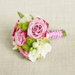 A small, romantic bouquet lies horizontally on a softly textured, light beige linen, as if just set down on a dining table in a Grove Park townhouse before a wedding. Two fully open pink roses form the heart of the design, their petals blending from a gentle blush at the centre to deeper magenta-pink along the outer edges, creating a subtle ombre effect that feels both modern and timeless. Between and around them, rounded tufts of bright apple-green button chrysanthemums nestle like tiny pom-poms, their many tightly packed florets adding playful texture and a lively contrast to the roses' velvety curves. Creamy white filler blooms, likely petite lisianthus or waxflower, thread through the arrangement, their starry centres and delicate petals softening the palette and giving the bouquet an airy, almost cloud-like lightness. Small touches of fresh green foliage peek through, grounding the colours in natural harmony. The stems are neatly trimmed and bound into a hand-tied posy, wrapped in a smooth, lustrous fuchsia-pink satin ribbon that catches the soft, even light. A fine strand of tiny white faux pearls spirals diagonally around the ribbon, like a bracelet, adding a hint of bridal jewellery to the handle. The neutral backdrop, with its visible woven texture, lets every shade of pink, green, and white stand out clearly, highlighting the careful craftsmanship and fresh, just-gathered feel. The overall mood is joyful yet refined, ideal for a bridesmaid's bouquet, a petite bridal posy, or a sweet centrepiece for an intimate celebration in a Grove Park home or local venue near the park itself, capturing the gentle excitement of a special day.