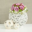 This carefully styled image shows a wedding table centerpiece that could grace a Grove Park riverside celebration, its gentle tones and textures arranged with an expert hand. At the centre stands a spherical white vase, its form created from slender, twig-like strands of rattan woven into an open, basket-like globe, where the spaces between the strands allow glimpses of shadow and subtle depth. Emerging from this sculptural base is a lush, domed bouquet composed mainly of roses in blush pink and soft lavender hues, some blooms displaying darker pink edging along the outer petals, adding a romantic, almost hand-painted effect. The roses are at full openness, revealing their layered, velvety petals in concentric spirals that catch the soft, even light used in the studio. Nestled among these larger blooms, clusters of bright green button chrysanthemums bring a cheerful, modern contrast, their compact, rounded shapes and fine texture reminiscent of the vibrant foliage found in nearby Grove Park gardens. At the lower edge of the arrangement, delicate white filler blossoms weave and spill gently over the rim of the vase, forming a light, cloud-like border that softens the structured dome and introduces a hint of natural looseness. The piece rests on a pristine white tabletop, the clean surface emphasising the arrangement's form and allowing the materials to stand out clearly. To the left of the vase, two decorative spheres crafted from pale wicker or rattan are placed in a relaxed grouping, one larger, one smaller, their open latticework echoing the vase's design and unifying the composition. The background is a softly blurred, light beige fabric with gentle texture, providing a warm, neutral setting so the flowers' colours and the crafted details remain the focus. The overall impression is of understated luxury, ideal for weddings and special occasions in Grove Park where elegance is expressed with calm, natural beauty.