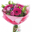 Against a clean, brilliant white background this hand-tied bouquet reads like a small celebration of spring: several large gerbera daisies in a vivid fuchsia-pink take centre stage, each with a very dark, almost black disc that gives the blooms a dramatic, velvety focus. Close to them nest a ring of delicate pink roses in varying stages of opening-some unfurled into soft, layered cups, others curled as tight, elegant buds-their pastel tones softening the gerberas' intensity. Dozens of glossy green leaves and slender stems form a verdant framework, the broad foliage giving the composition depth and a fresh, garden-born feel. Interspersed among the roses and gerberas are small clusters of deep blue-purple statice (or similar filler flowers), adding fine, papery texture and tiny points of cool colour. The bouquet is wrapped in layers of crinkled, sheer pink cellophane that graduates from an almost translucent pastel halo at the top to a richer, more saturated pink at the base, catching soft studio light and creating a gentle bloom of colour around the heads. The bundled stems sit inside a clear cylindrical glass vase so the shuttered, water-kept stalks are visible; a piece of rustic twine is tied around the vase neck and a small white rectangular tag bearing a stylised pink and green rose logo hangs like a quiet signature. I picture this arrangement as a cheerful gift for a birthday, a Mother's Day surprise, or to brighten a local table in Grove Park or nearby Lee; the scent is lightly sweet and floral, the textures velvety and crisp - a bouquet that feels both carefully crafted and naturally generous.