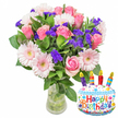 Bathed in soft, even light against a clean white backdrop, this celebratory arrangement reads as a bright, tactile gift: a lush bouquet of freshly cut blooms set in a clear glass vase with green stems and water visible at the base, paired with a playful cake-shaped foil balloon. The flower composition centers on fully opened soft-pink roses, their velvety petals unfurling from pale blush to a warmer, more saturated pink at the hearts, while generous pale-pink Gerbera daisies-each with layered, ray-like petals and buttercream-yellow centers-form cheerful focal points that feel almost like smiling faces. Woven between the larger blossoms are clusters of vivid purple filler flowers, likely statice or limonium, whose crisp, papery texture gives depth and a jewel-like contrast to the pinks. Small white spray blooms add flecks of brightness while glossy, deep-green foliage frames and supports the whole, giving the bouquet a natural, freshly gathered look.
To the right, a white, cake-shaped Mylar balloon stands as a festive partner: a frosted surface trimmed with a wavy blue icing border, scattered multi-colored sprinkles in red, yellow, blue and green, and four striped candles-purple, red, orange and blue-each topped with a tiny yellow flame. The words Happy Birthday appear across the front in playful, multi-colored type and a small KALEIDOSCOPE logo sits at the balloon's base. There is a gentle, sweet fragrance suggested by the roses and greens, and the overall impression is joyous and affectionate-an ideal birthday gesture from a florist who values texture, colour and freshness.