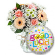 A bright, encouraging 'Good Luck' gift captured against a clean white background: to the left a generous, hand-arranged bouquet sits in a clear glass vase tied simply with rustic twine, the light catching the vase's rim and the soft sheen of the stems. The arrangement combines soft peach-orange Gerbera daisies with their dark brown centres and romantic, tightly furled pale pink roses; tiny clouds of white baby's breath (Gypsophila) are woven through the blooms like lace, while small white and lime-green chrysanthemum florets add crisp contrast. Deep green foliage peeks between the blossoms, giving the bouquet a lush, grounded silhouette and a subtle leafy scent beneath the rose perfume. In the foreground and overlapping the lower right of the vase is a round metallic silver foil balloon, its surface gleaming with small, colorful stars, hearts and confetti shapes. The playful message Good Luck! appears in chunky, multi-coloured letters - the two o's in Good replaced by cheerful yellow smiley faces, one winking, one making a tongue-out expression - and a tiny KALEIDOSCOPE logo sits low on the balloon's edge. The composition is lit with soft, even studio light that emphasises pastel tones and the balloon's metallic sparkle. This pairing feels hopeful and reassuring: the tactile velvet of rose petals, the papery crispness of gerbera petals and the cool, smooth sheen of the Mylar balloon combine to create a comforting, celebratory gift ideal for someone in Grove Park setting out on exams, interviews, farewells or a new beginning.
