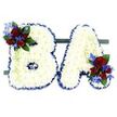 A solemn, handcrafted tribute in the shape of the capital letters 'B' and 'A', carefully formed from hundreds of small, pristine white chrysanthemum heads that create a cloud-like, pillowy surface. Each letter's contours are crisply defined by a ruffled royal blue satin ribbon that traces the outer edge, giving a quiet but dignified contrast against the pure white blooms. The 'B' carries a compact cluster of deep red roses on its upper left curve - roses at different stages from tight buds to gently opened cups - which are punctuated by vivid, spiky blue delphinium-like florets and softened with slender, pale green trailing foliage and darker glossy leaves. The 'A' balances this composition with a matching cluster tucked into its lower right, repeating the same palette of velvety red, electric blue, and lush green so the pair read as one cohesive memorial. Both letters sit side-by-side on a low, dark green rectangular base that keeps the arrangement steady and subtly grounds the composition against the bright white background, which allows the colours and textures to read clearly in soft daylight. The overall effect is one of composed respect and quiet reverence - a personalised floral initial tribute often chosen for funerals or memorials to convey remembrance and love. As a florist who has placed similar pieces at Grove Park station chapels and local memorial services near Chinbrook Meadows, I notice how the cool white chrysanthemums seem to glow under morning light, lending the tribute a sense of peace and lasting care.
