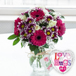 A vibrant bouquet overflowing with colour stands in a simple, rounded glass vase on a light, clean surface, caught in gentle daylight reminiscent of a bright morning in Grove Park. At the centre, bold gerbera daisies unfurl in deep fuchsia and magenta, their large, daisy-like faces completely open and anchored by rich, dark centres that give the arrangement a strong, modern focus. Around them, compact clusters of chrysanthemums provide a striking contrast, each blossom showing a ring of deep purple that fades to crisp white at the tips of the petals, circling a lively yellow core that adds a spark of cheer. Creamy white roses nestle among these richer hues, their soft, layered petals offering a sense of calm and subtle romance amid the bolder colours. Interlaced throughout are delicate fronds of white baby's breath, creating a fine, cloud-like texture that makes the bouquet feel both abundant and airy. Glossy green foliage peeks out from beneath and between the blooms, echoing the leafy character of the local streets and riverside walks around Grove Park. The stems of the flowers are clearly visible through the transparent vase, neatly trimmed and submerged in clear water, highlighting the freshness of the arrangement. A length of rustic, natural twine is tied securely around the neck of the vase, and from it dangles a small, white tag bearing a stylised pink rose and friendly green script, suggesting a gift thoughtfully prepared by a local florist. To the right, a heart-shaped foil balloon leans into the frame, outlined in reflective silver. Its white centre is printed with colourful, mosaic-style lettering in shades of pink, purple, and blue, spelling out the affectionate phrase Love n' Hugs 24-7, accented with tiny hearts that underscore the loving theme. Set against a softly blurred, luminous background, this combination of flowers and balloon radiates warmth and celebration, ideal for a birthday, anniversary, or just-because flower delivery anywhere in Grove Park.