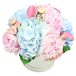 Softly opulent pastel bouquet arranged in a round white hat box, set against a spotless white background that lets every petal and colour nuance stand out. At the heart of the design, large pink peonies bloom in layers of delicate, frilled petals, some wide open and others still cupping their centres, creating a plush, romantic focal point. Surrounding these are generous clusters of light blue hydrangeas, their countless small florets combining into cloudlike spheres that add depth, volume, and a cool, soothing tone. Interwoven between the bigger blossoms are dainty light pink spray roses, many still in tight bud, others just beginning to reveal their inner petals, contributing intricate detail and a sense of gentle anticipation. Lifting above the rounded mound of pastels, two vivid pink calla lilies curve with sculptural elegance, one placed slightly to the left and the other to the right, their sleek, trumpet-shaped blooms bringing a modern, refined touch to the composition. A few deep green leaves peek out discreetly at the base, offering a quiet hint of foliage that anchors the palette without overpowering it. The flowers fill the smooth white hat box so generously that they appear to overflow, while a subtle ridge around the top edge adds a small decorative note to the container. Soft, even lighting enhances the velvety textures and makes the colours glow softly rather than shout. This beautifully balanced arrangement evokes the calm of a walk along the river at Grove Park, and would feel perfectly at home as a baby welcome gift, a graceful sympathy tribute, or a centrepiece for a spring gathering in a house near Kew Bridge or Strand-on-the-Green.