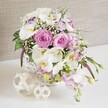 An abundant floral arrangement in a round white wicker basket is presented like a centerpiece ready for a wedding table in Grove Park, its colours and textures captured in soft, diffused light. The design is dominated by large, fully open pink roses, their petals curving out in perfect spirals that speak of romance and celebration, as if created for a special gathering near Grove Park station or along the Thames path. Interlaced with the roses are elegant white Phalaenopsis orchids, their broad petals smooth and slightly curved, encircling golden-yellow centers that add a subtle glow. White Lisianthus blossoms, some with delicate lavender-purple outlining their ruffled edges, rest between the larger flowers, offering a gentle, layered softness. Pale pink stock stems rise and fall over the edges of the basket, suggesting a slight cascading form that adds fluidity, like a bridal bouquet reimagined as a table arrangement. Tiny sprigs of baby's breath are sprinkled throughout, forming airy white clusters that lighten the overall effect and soften the transition between blooms. Rich green foliage peeks in and out, some leaves tucked deep among the flowers, others subtly tumbling over the basket's rim, giving the composition a fresh, garden-inspired look. The white wicker basket itself, with its neatly woven pattern, stands on a clean white surface. Nearby, two decorative spheres crafted from fine strands of rattan or wicker echo the basket's texture and introduce a contemporary, handcrafted note often seen in stylish Grove Park interiors. Behind, a warm beige, linen-like background keeps the focus firmly on the subtle interplay of pinks, whites and greens. The scene conveys a mood of gentle elegance and joyful celebration, perfect for weddings or refined table settings throughout the Grove Park area.