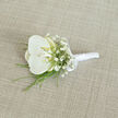 Resting at a gentle angle on a pale, woven fabric that looks like finely textured linen in soft beige, this elegant boutonnière is captured in calm, even light that reveals every careful detail. The centrepiece is a single, immaculate white orchid bloom, its wide, silky petals curving outwards with effortless grace, like a small, sculpted piece of porcelain. At the heart of the orchid, the throat glows with a muted touch of buttery cream and subtle yellow, introducing a delicate warmth that softens the coolness of the white. Surrounding the central flower, airy sprays of white baby's breath are tucked in with precision, their numerous tiny blooms creating a filigree of texture that suggests lightness, purity, and a whisper of romance. Emerging from the base, fine strands of vivid green foliage, perhaps ornamental grass or fine fern tips, arc gently outwards, adding a slender frame of colour that echoes the fresh greenery seen in Grove Park's gardens and tree-lined avenues. The stems of the flowers are tightly gathered and wrapped from base to head in smooth white satin ribbon, the fabric gleaming softly and forming a clean, cylindrical handle designed to sit comfortably on a lapel throughout the day. Near the top of the ribbon, just under the orchid, a neatly tied satin bow adds a tailored finishing touch, suggesting careful hands and an experienced florist's eye. The entire composition has a quiet, sophisticated charm, ideal for a groom or groomsman heading to a wedding near Grove Park or along the Thames, conveying understated luxury, freshness, and a sense of poised anticipation for the vows and celebrations to come.
