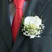 A detailed close-up shows the upper torso of a person dressed for a special celebration in Grove Park, captured just as they adjust their jacket before stepping out. The dark suit, a rich charcoal grey that almost reads as black, is sharply tailored and framed so that the right lapel fills much of the image. Sitting proudly on that lapel is a carefully crafted boutonniere centred around a single white Phalaenopsis orchid. Its broad, velvety petals curve outward in a soft, luminous arc, with a creamy yellow throat that seems to glow gently against the darker fabric. Around the orchid, clusters of tiny white baby's breath form a delicate cloud, their miniature blossoms adding fine texture and a sense of lightness. Slender blades of bright green, grass-like foliage thread through the arrangement, providing movement and a fresh, garden-inspired touch that feels at home in a leafy area like Grove Park. The stems are neatly bound and finished with a narrow white sheer ribbon, tied into a small, precise bow that keeps the design elegant rather than showy. Beneath the flowers, a lustrous red silk tie cuts diagonally through the composition, its smooth surface catching the light and contrasting boldly with a subtly patterned, pale dress shirt. The overall mood is one of poised anticipation-a groom or prom guest ready for photographs on a tree-lined street near the station, the cool, fresh petals against the lapel a quiet reminder of the care and craftsmanship that goes into every formal floral detail for weddings and evening events in this part of London.
