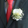 A single white orchid, luminous and composed, rests above a suit pocket like a small promise on a wedding morning in Grove Park. Its petals are softly rounded and creamy, catching the light with a subtle sheen that lends them a porcelain-like quality. At the centre of the bloom, a flicker of pale gold glows, a hidden warmth that draws the eye closer. Wrapping around this focal flower, clusters of baby's breath form a delicate halo, each tiny white blossom adding a sense of lightness and air. Fine green stems and leaves weave between the gypsophila, introducing a touch of natural movement and echoing the leafy calm of the Grove Park area and the nearby Thames path. The boutonnière is bound at the base with a slim white ribbon, tied into a neat, understated bow that suggests experienced hands and careful attention to detail. The flowers sit against a deep charcoal lapel, whose matte texture throws the crisp whites and greens into sharper relief. Alongside, a striking red tie creates a bold sweep of colour, the rich, saturated crimson bringing energy and romance to the composition. Together, these elements form an image that feels both classic and contemporary, perfect for a groom or best man standing outside a Grove Park church or town hall. The interplay of gentle curves and sharp tailoring, soft petals and strong fabric, speaks of balance and harmony. It is a boutonnière that does not shout, but rather quietly holds the emotion of the day-love, commitment, and the significance of marking life's milestones with something beautiful and thoughtfully made.