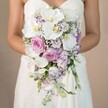 Nestled in the gentle light typical of a Grove Park morning, this photograph features a bride in an elegant, strapless lace gown, shown holding a luxuriously cascading bridal bouquet that falls gracefully from her hands. The bouquet's foundation is a cluster of pristine white phalaenopsis orchids, their creamy petals framing vivid yellow centers. Soft pink garden roses contribute layers of romantic texture, while lavender stock and light purple lisianthus-some fluttering with a hint of deep violet on their edges-are scattered among the larger blooms. Wispy baby's breath drifts lightly through the bouquet, adding a delicate mistiness reminiscent of riverside walks near Grove Park. Minimal but intentional greenery enhances the organic aesthetic without detracting from the floral display. The overall palette-soft purples, gentle pinks, and timeless whites-creates a visual harmony that would complement any wedding in Grove Park. The image exudes refined elegance, artisan expertise, and the warmth infused into every arrangement by the community's talented local florists.