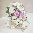 A refined, contemporary floral display sits serenely on a white tabletop, bathed in soft light much like an elegantly set room near Grove Park station on a wedding morning. Several Phalaenopsis orchids in pristine white rise gracefully from the centre, their broad, smooth petals curving in arcs that feel both delicate and poised. Each orchid reveals a golden-yellow and muted orange throat, a warm focal point that draws the eye inward. Between these sculptural blooms nestle lavender-pink roses, fully open, their spiral petals arranged in tight, luxurious layers that invite a sense of closeness and affection. Wisps of white baby's breath are woven into the arrangement, their tiny blossoms forming a gossamer veil that gently links one flower to another. On the outer edges, light purple lisianthus, some brushed with deeper violet along their frilled rims, pair with cascading stems of small pastel lavender-pink flowers that fall softly over the side, gifting the bouquet a gentle, cascading rhythm. A frame of lush green leaves, including broad, glossy shapes and finer textures, supports and contrasts the softer tones, lending the display depth and structure. The flowers are gathered in a striking white vase made of narrow interlaced strips, forming an open, spherical lattice that feels both modern and organic. At the lower left, two decorative woven spheres, one larger, one smaller, rest beside the vase, echoing its pale, openwork design and adding visual balance. The background is a softly lit beige surface with subtle texture, enhancing the sense of calm and refined taste. This arrangement conveys quiet joy and enduring love, perfect for weddings, anniversaries, or elegant celebrations across Grove Park and the surrounding Chiswick area.
